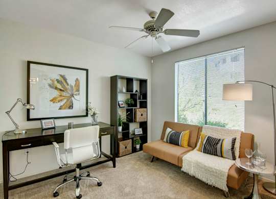 A versatile home office and guest space here at Mirador featuring soft neutral walls, plush carpeting, and a ceiling fan overhead. A sleek dark wood desk with a modern chair sits beneath framed artwork, while a cozy sofa with accent pillows and a throw rests beside a floor lamp and large window with blinds that invite natural light into the room.