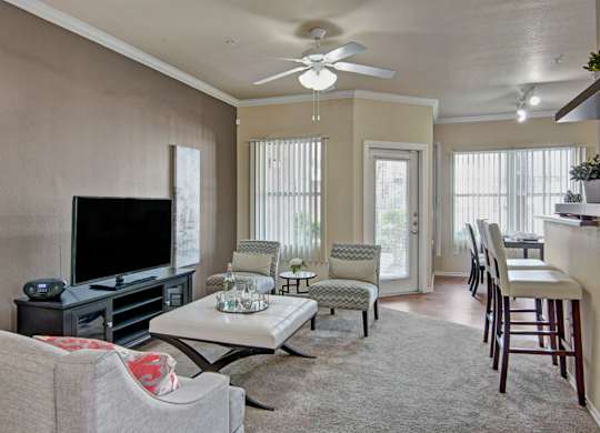 A bright and inviting living room here at Mozaic at Steele Park featuring plush carpet, neutral walls, and a ceiling fan with light. Large windows with vertical blinds and a glass door bring in natural light, while the open layout flows into a kitchen with bar seating, creating a comfortable space for relaxing or entertaining.