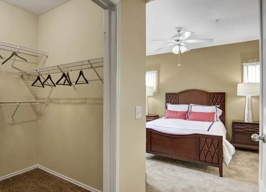 A spacious bedroom with walk-in closet here at Mozaic at Steele Park featuring plush carpet, neutral walls, and a ceiling fan with light. The open closet includes built-in shelving and hanging space, while the adjoining bedroom comfortably fits a full bedroom set with matching nightstands for added storage.