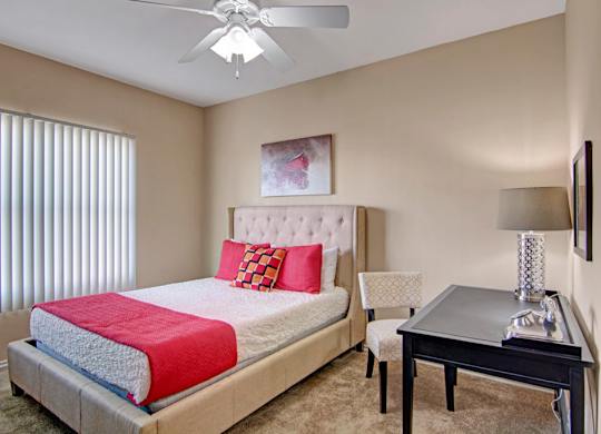 A cozy bedroom here at Mozaic at Steele Park featuring plush carpet, neutral walls, and a ceiling fan with light. A large window with vertical blinds provides natural light, while the spacious layout accommodates a bed, nightstand, and desk area, offering both comfort and functionality.
