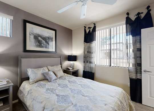 A serene bedroom here at Natura Villas featuring a plush bed with patterned bedding and coordinating accent pillows, framed by soft neutral walls and a contrasting accent wall. Matching bedside tables with modern lamps add balance, while large windows with vertical blinds and layered curtains invite natural light, creating a calm and comfortable retreat.