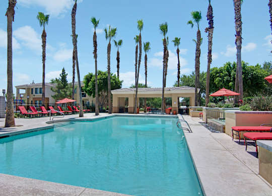 A resort-style swimming pool here at Natura Villas surrounded by tall palm trees and expansive sun decks, featuring crystal-clear water, multiple lounge chairs with vibrant red cushions, and shaded seating areas under umbrellas. A covered pavilion offers additional gathering space, creating a relaxing outdoor retreat perfect for enjoying sunny Arizona days.