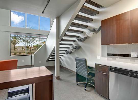 A modern loft-style living space here at Proxy 333 featuring dramatic open-riser stairs with wood treads, tall windows bringing in natural light, and polished concrete floors, complemented by a sleek kitchen with dark cabinetry, a built-in workspace, and a dining island that enhances the airy, contemporary layout.
