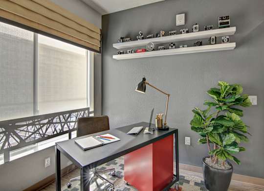 A stylish home office nook here at Proxy 333 featuring a modern desk with a task lamp, rolling chair, and workspace accessories, set against a gray accent wall with floating shelves displaying vintage cameras, complemented by a large window with a shade for natural light and a leafy floor plant adding warmth.