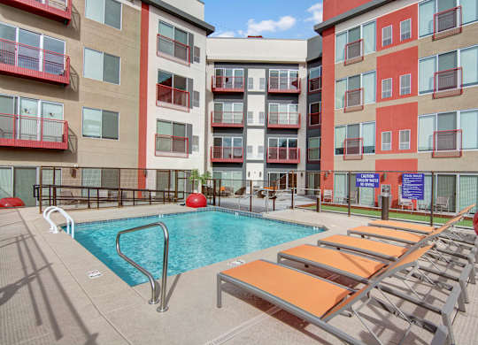 A modern outdoor pool courtyard here at Proxy 333 featuring a rectangular swimming pool with stainless handrails, surrounded by orange lounge chairs on a spacious concrete deck, set against contemporary apartment buildings with red-accented balconies, secure fencing, and shared seating areas creating a vibrant urban retreat.