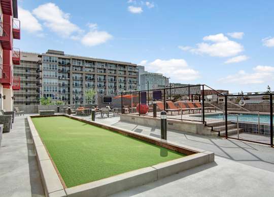 A spacious outdoor amenity deck here at Proxy 333 featuring a vibrant green turf game area bordered by concrete walkways, overlooking a fenced swimming pool with lounge seating, orange accents, and city views in the background, all set beneath an open blue sky that creates a bright and inviting urban atmosphere.