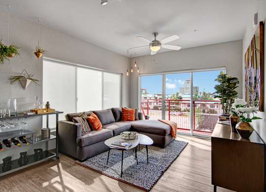 A bright contemporary living room here at Proxy 333 showcasing wood-style flooring, a spacious gray sectional with warm accent pillows, and a textured area rug anchored by nesting coffee tables, while large sliding glass doors open to a private balcony and city views, complemented by a ceiling fan, modern lighting, decorative plants, and stylish wall accents.