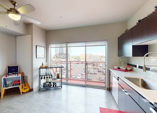 A bright open living space here at Proxy 333 featuring wood-style flooring, sleek dark cabinetry with a modern tile backsplash, and a streamlined kitchen with a large sink and expansive countertops, flowing into a flexible living area with a ceiling fan, bar cart, and decorative accents, while sliding glass doors open to a private balcony with city views and abundant natural light.