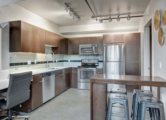 A contemporary kitchen here at Proxy 333 featuring sleek dark wood cabinetry, a light tile backsplash, and smooth countertops paired with stainless steel appliances, complemented by track lighting, a modern breakfast bar with seating, and polished concrete-style flooring that creates a clean, open, and functional cooking and dining space.