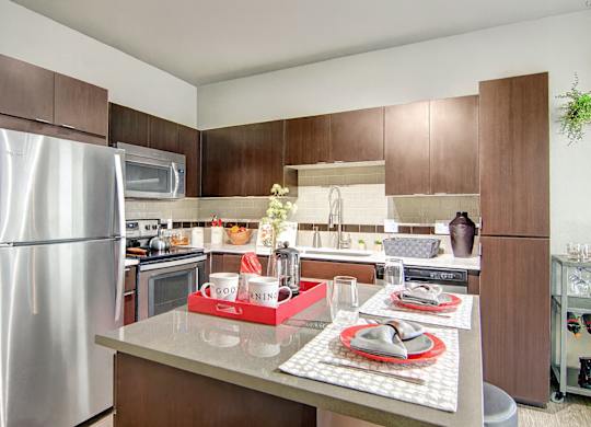 A modern kitchen here at Proxy 333 showcasing rich dark wood cabinetry, smooth light countertops, and a spacious island set with place settings, complemented by stainless steel appliances, a tiled backsplash, undercabinet lighting, and clean contemporary finishes that create a polished and inviting cooking and dining space.