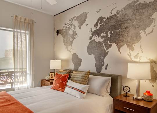 A stylish bedroom here at Proxy 333 featuring a comfortable bed dressed in crisp neutral bedding with layered accent pillows, matching nightstands with modern table lamps, a striking world map accent wall, soft window coverings, and a large window that brings in natural light for a warm and inviting retreat.