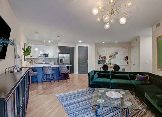 A stylish open living room here at Roosevelt Row showcasing a plush emerald sectional sofa with accent pillows, a glass coffee table set on a patterned area rug, and wood-style flooring flowing into a modern kitchen with an island, bar seating, sleek cabinetry, and stainless-steel appliances, all accented by contemporary artwork and statement lighting.