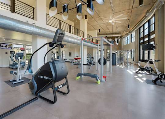 A spacious fitness center here at Roosevelt Row showcasing an open-concept gym with high ceilings, exposed ductwork, large windows for natural light, and a variety of cardio machines, strength equipment, and free weights arranged across polished flooring, creating a modern and motivating workout environment.