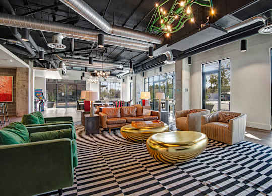 A spacious community lounge living room here at Roosevelt Row featuring an open industrial design with exposed ductwork, high ceilings, and large windows that fill the space with natural light, highlighted by striped area rugs, plush sofas and armchairs, sculptural metallic coffee tables, modern lighting fixtures, and inviting seating areas ideal for socializing and relaxing.