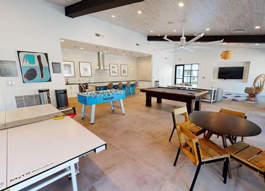 A lively community game lounge living room here at Roosevelt Row showcasing an open, airy layout with high ceilings, exposed beams, and modern ceiling fans, featuring a ping pong table, foosball table, and pool table alongside a long bar with seating, framed artwork, tiled flooring, and cozy seating areas designed for socializing, games, and resident gatherings.