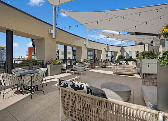 A spacious rooftop terrace here at Roosevelt Row featuring woven lounge seating with patterned cushions, café-style tables and chairs, large planters with greenery, and shade sails strung overhead with café lights, all set on tiled flooring with city views visible beyond the open railing, creating a relaxed outdoor gathering space for residents.