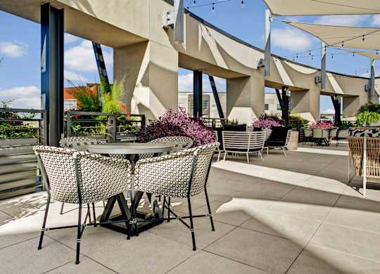 A sunlit rooftop patio here at Roosevelt Row showcasing café-style round tables with woven patterned chairs, sleek concrete pavers, and modern architectural columns, surrounded by raised planters filled with colorful greenery, string lighting overhead, and shaded seating areas that create an inviting space for relaxing, dining, or socializing with city views.