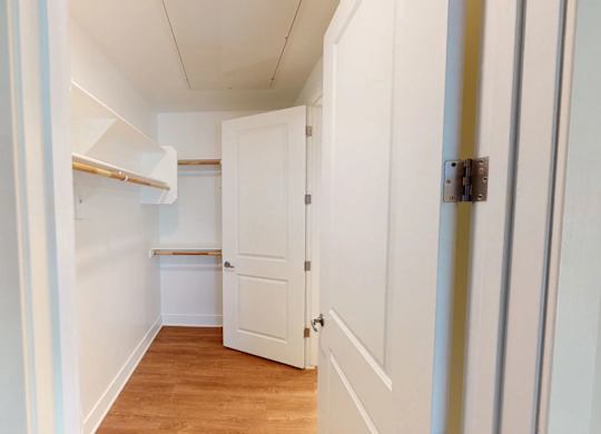 A spacious walk-in closet here at Roosevelt Row featuring crisp white walls, built-in shelving with upper storage cubbies, dual hanging rods for organized clothing space, wood-style flooring, and a clean, open layout that offers practical storage and easy access through paneled doors.