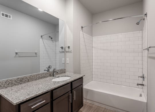 Modern bathroom here at 49 West Apartments featuring a large mirror, granite countertop, and dark cabinets. A bathtub with white subway tiles and chrome fixtures adds elegance.