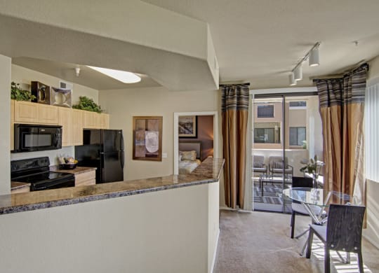 Modern kitchen and dining area here at Natura Villas Apartments with light wood cabinets, black appliances, and a marble counter. A glass table with chairs sits by wide patio doors.