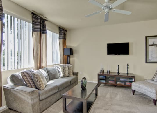 Bright, modern living room here at Natura Villas Apartments with a gray sofa, patterned pillows, and a sleek coffee table. Sunlight streams in through vertical blinds, creating a calm ambiance.