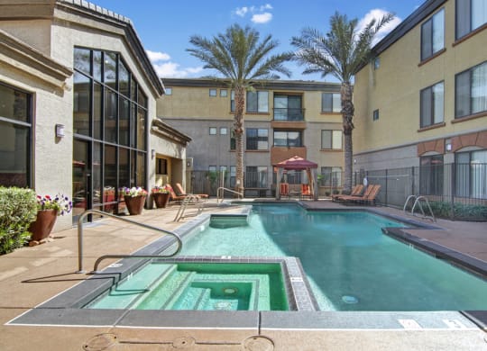 Outdoor pool and hot tub area here at Bolero Apartments surrounded by loungers, palm trees, and beige buildings under a bright blue sky with fluffy clouds. Relaxing ambiance.