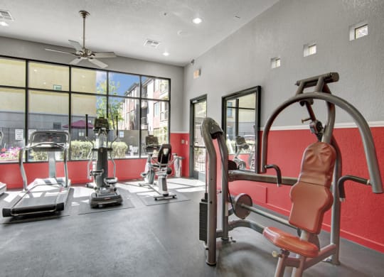 Bright gym here at Bolero Apartments with large windows showing a sunny outdoor view. Room includes treadmills and a stationary machine on red and gray walls, conveying energy.