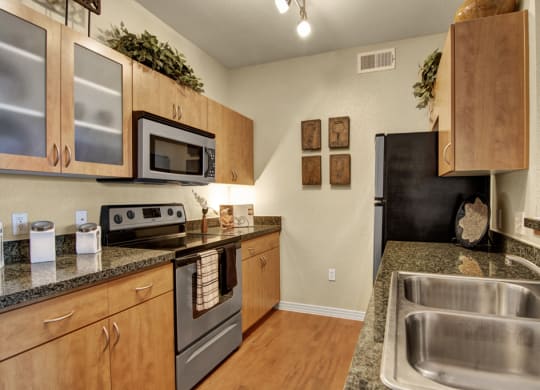 A cozy kitchen here at Bolero Apartments with wooden cabinets and granite countertops. It features a stainless steel stove, microwave, and a double sink. Decorative plants and art add warmth.