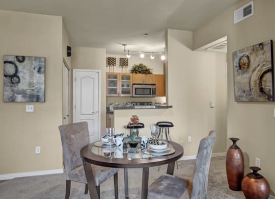 A cozy dining area here at Bolero Apartments with a round table set for two, featuring elegant dishware. Abstract art adorns beige walls, and a warm kitchen is visible in the background.