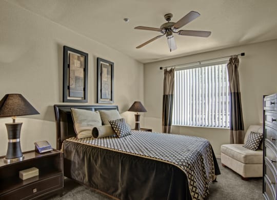 Cozy bedroom here at Bolero Apartments with neutral tones, featuring a large bed with patterned linens, two bedside lamps, wall art, a window with blinds, and a plush chair.