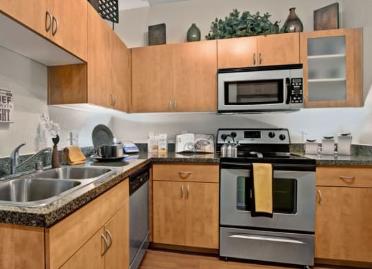 Modern kitchen here at Bolero Apartments with wooden cabinets, granite countertops, stainless steel appliances including a stove and microwave. A yellow towel hangs from the oven, and decorative plants sit above the cabinets, adding a cozy feel.