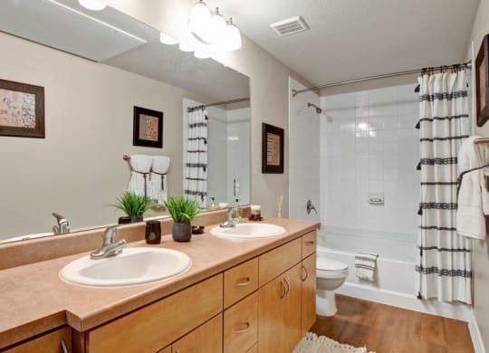 Modern bathroom here at Bolero Apartments with double sinks on a wooden vanity, large mirror, and white tiled shower with striped curtain. Decor includes framed art and plants.