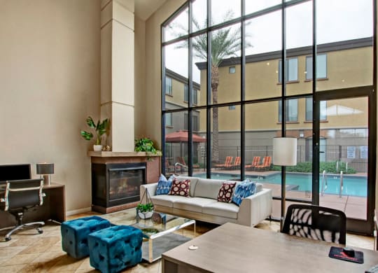 Spacious modern lobby here at Bolero Apartments with high glass windows overlooking a pool area. Features cozy seating, a fireplace, vibrant blue stools, and computer desks.