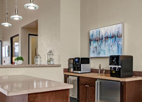 Modern office breakroom here at Bolero Apartments with a coffee station, soft lighting, abstract wall art, and a stone countertop. The ambiance is clean and inviting.