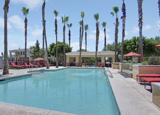 Sunny outdoor pool area here at Natura Villas Apartments with tall palm trees and red lounge chairs. A playground is visible to the left, and a shaded pavilion is at the pool's end.