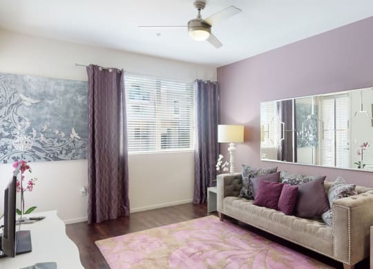Modern living room here at Artisan at Main Street Metro Apartments with lavender walls, grey tufted sofa, and pink cushions. Large mirror, abstract art, and floral rug complement the chic decor.