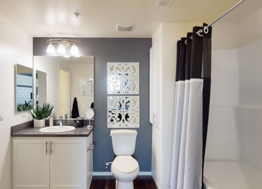 A modern bathroom here at Artisan at Main Street Metro Apartments with a sleek design features a white vanity, round basin, and rectangular mirror. There's a toilet, decorative wall art, and a shower with a black and white curtain. The space is bright and organized.