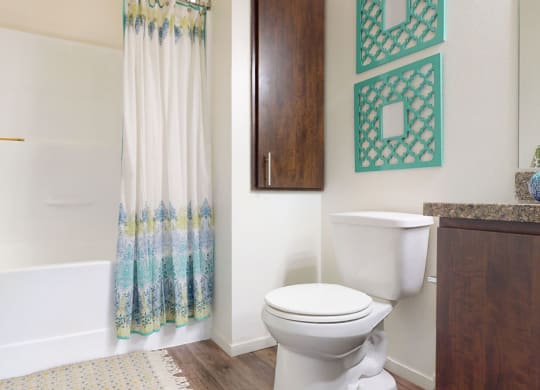 Bright bathroom here at Artisan at Main Street Metro Apartments with a white tub and shower, blue-patterned curtain, wood cabinet, white toilet, and turquoise wall decor. Cozy and clean atmosphere.
