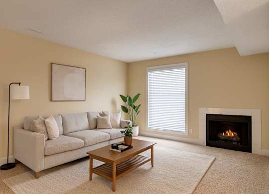 A cozy living room here at 3600 Brentwood with soft beige carpet, a light gray sofa, a wooden coffee table, and a warm fireplace, accented by a bright window with white blinds and simple modern decor.