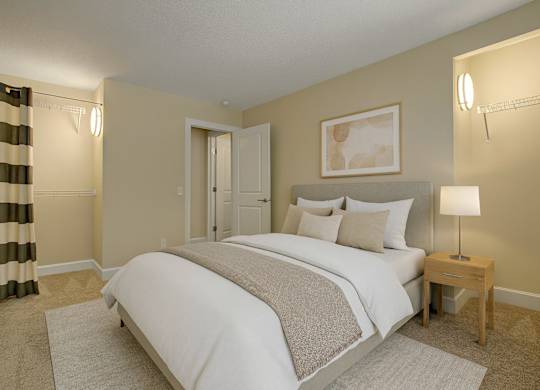 A cozy bedroom here at 3600 Brentwood with soft neutral carpeting, a comfortable bed with crisp white bedding, and warm accent lighting, featuring open closet storage and a clean, modern feel.