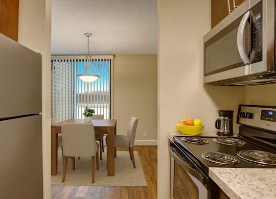 A modern kitchen and dining nook here at 3600 Brentwood with stainless steel appliances, a sleek stove, and a cozy dining table set beside tall windows with vertical blinds for natural light.
