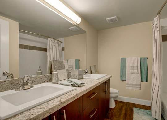 A modern bathroom here at 3600 Brentwood with a double-sink vanity, granite-style countertops, and a large mirror, featuring a bathtub-shower combo and clean, bright lighting.