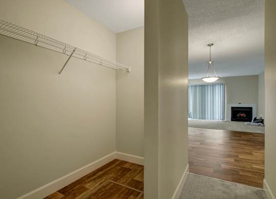 A convenient walk-in closet here at 3600 Brentwood with open wire shelving and warm wood-style flooring, leading into the bright living area with a dining light and cozy fireplace in the distance.