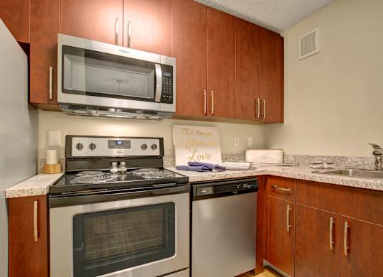 A modern kitchen here at 3600 Brentwood with rich wood cabinetry, granite-style countertops, and stainless steel appliances including a microwave, dishwasher, and stove, plus a sleek sink for everyday convenience.