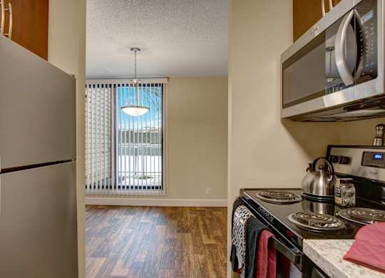 A modern kitchen here at 3600 Brentwood with stainless steel appliances, an electric stove, and granite-style countertops, opening into a bright dining area with wood-style flooring and tall windows with vertical blinds.