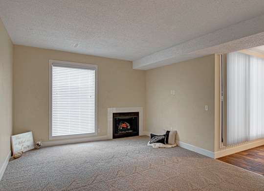 A spacious, open living room here at 3600 Brentwood with soft carpet, a cozy fireplace, and tall windows with blinds that bring in natural light, flowing into the dining area with wood-style flooring.