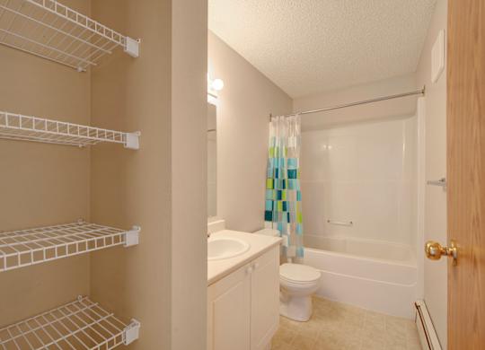 A clean bathroom here at Aspen Terrace with a bathtub-shower combo, a white vanity with a smooth countertop, and a bright mirror light. A built-in shelving nook with wire racks provides extra storage, giving the space a practical, fresh feel.