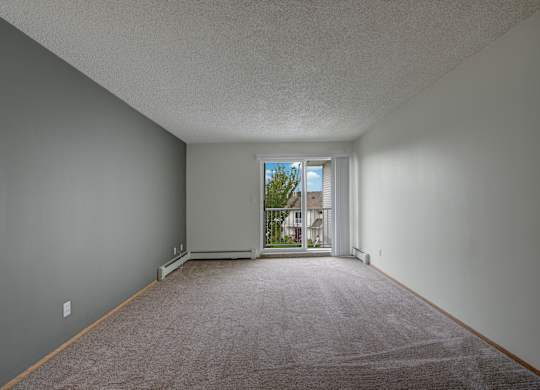 A spacious, empty living room here at Aspen Terrace with soft carpet, neutral walls, and a sliding glass door that opens to a private balcony. A large window and bright natural light create an airy, inviting feel.