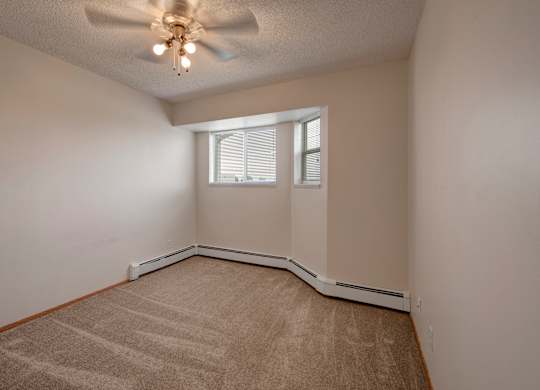 A bright, empty bedroom here at Aspen Terrace with beige carpet, neutral walls, and a ceiling fan with overhead lighting. A corner window with blinds brings in natural light, creating a cozy, comfortable feel.