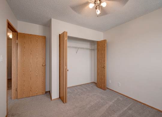 A bright, empty bedroom here at Aspen Terrace with soft carpet, neutral walls, and a ceiling fan with overhead lighting. A roomy closet with wood doors provides convenient storage, creating a clean, comfortable feel.